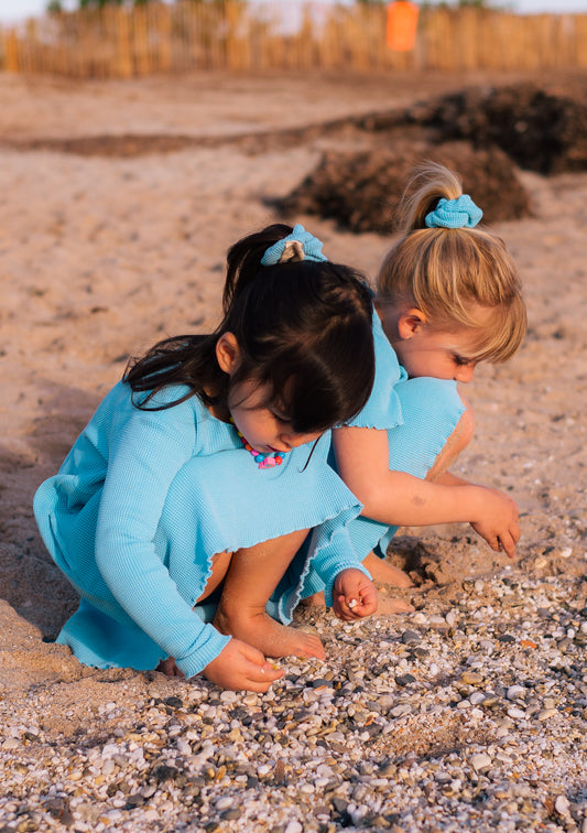 A toddle standing by the ocean and looking at sunset. She is wearing a beautiful organic cotton yellow dress with long sleeves and pockets.