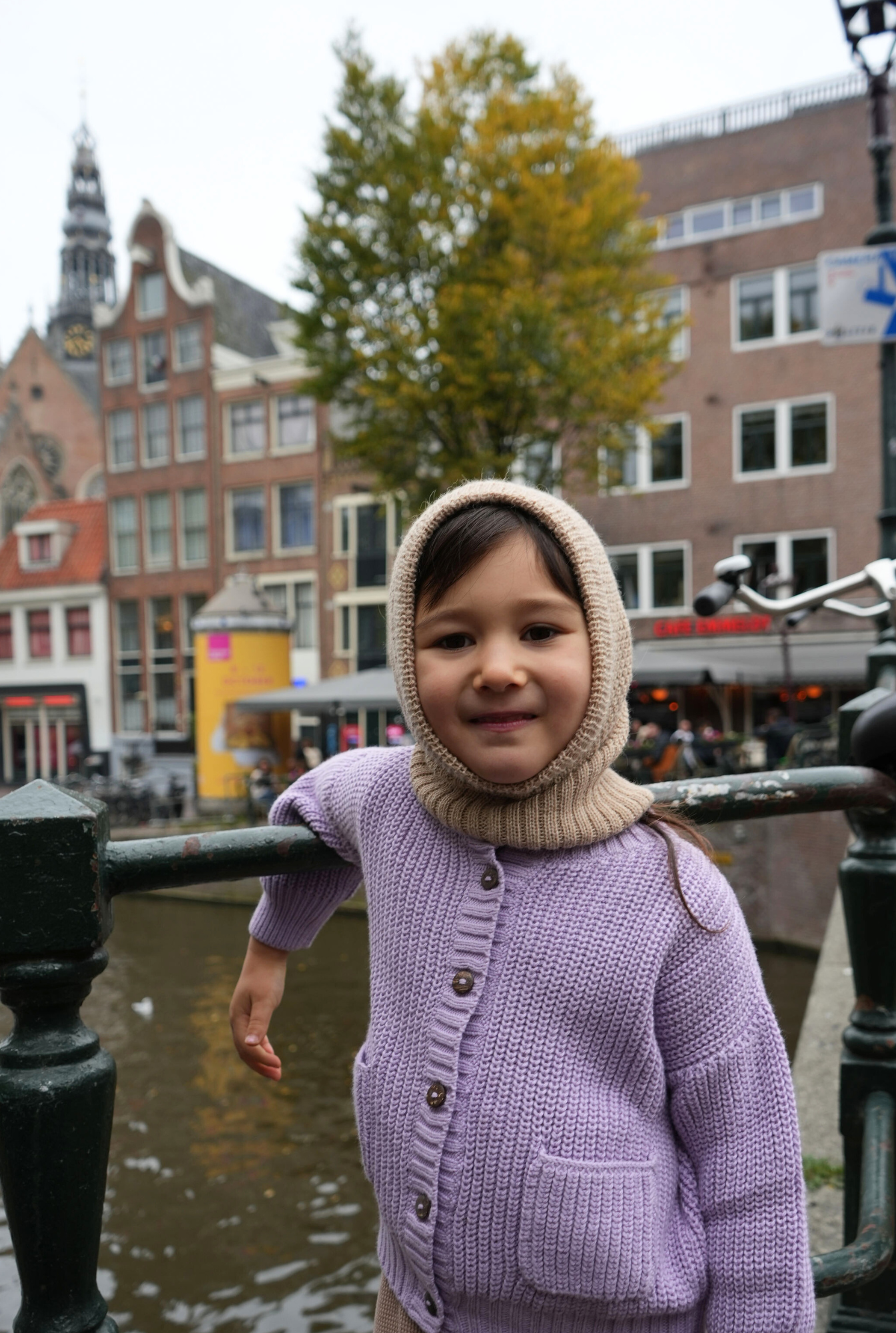 Child in a purple 100% tanguis cotton sweater and beige balaclava hat made with baby alpaca standing by a canal with historic buildings in the background.