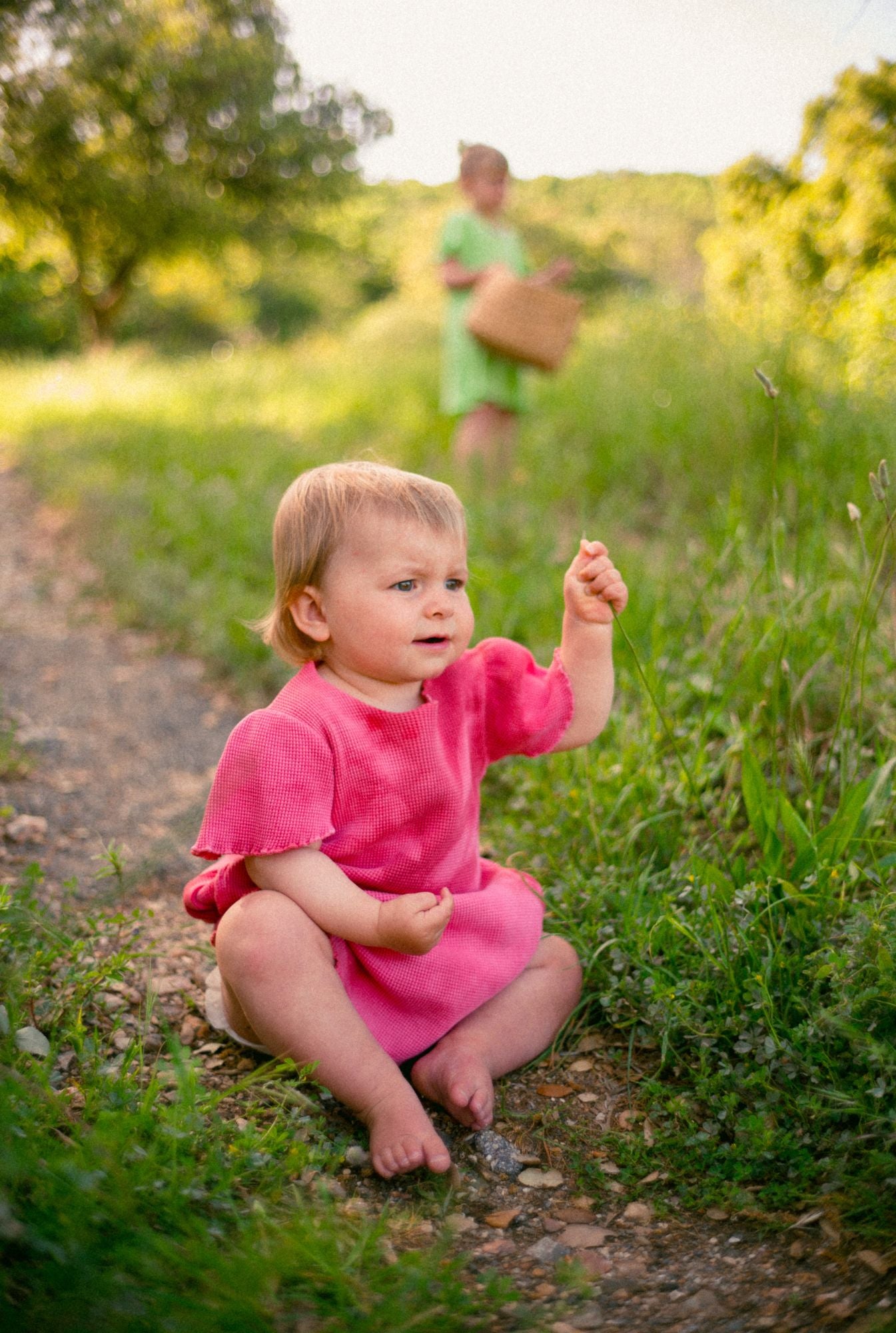 1 year old girl is playing with flowers and wearing a beautiful, comfortable, and colorful dress in pink tie dye. The dress is made by Tiny Advocates. It comes in a variety of beautiful colors, has side pockets, and beautiful details around the neckline and sleeves.