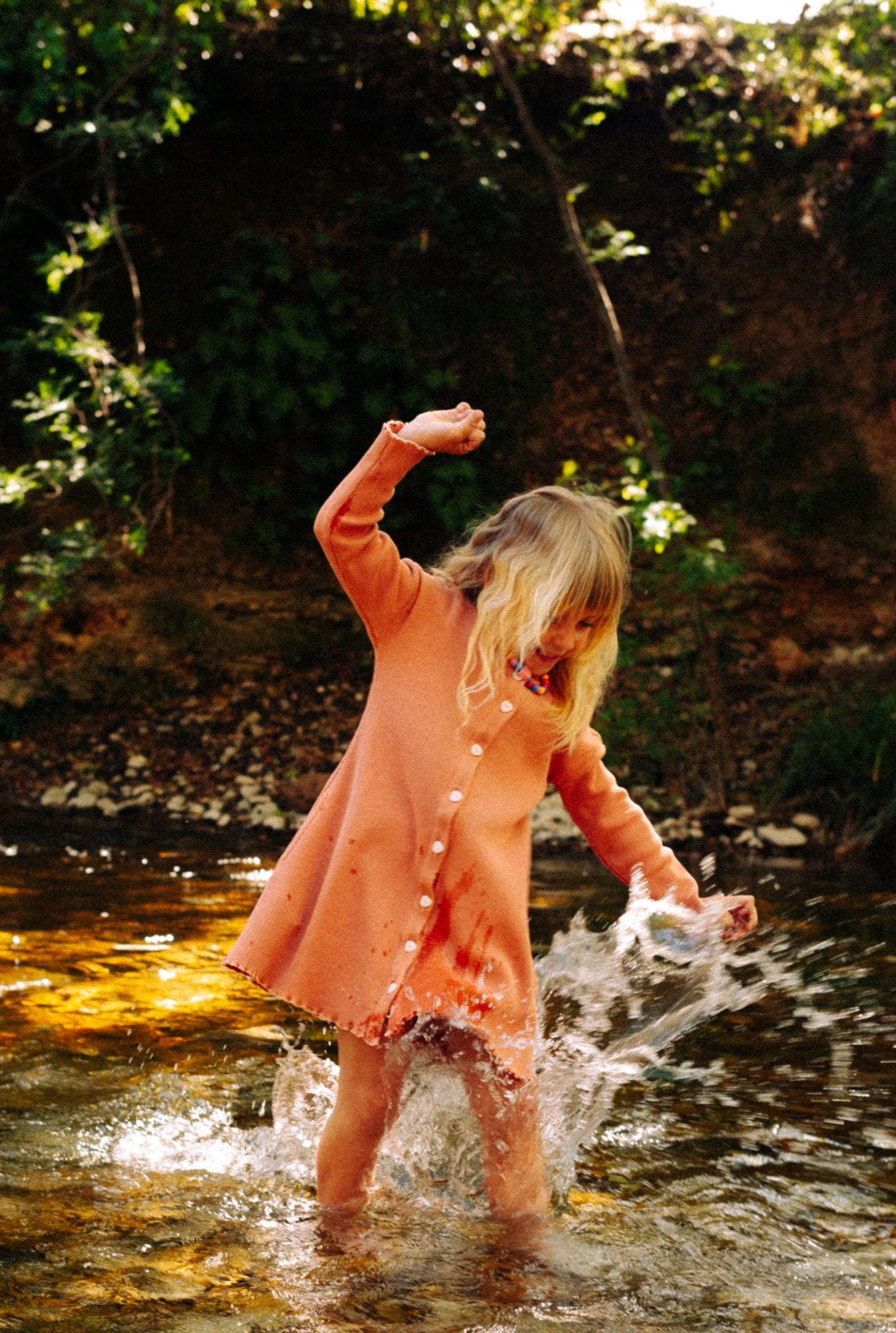A little girl playing in the river getting splashed by water. She is wearing a comfortable dusty rose dress. The dress is made from 100% organic cotton and garment dyed in natural minerals. It is breathable, comfortable, and fun for everyday play.