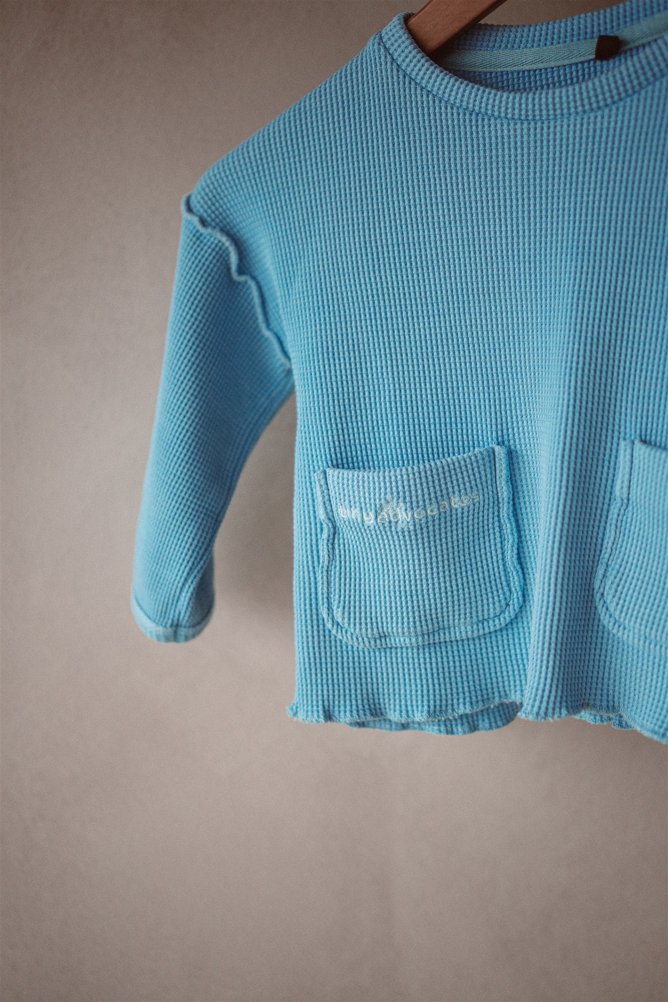 An image of organic cotton kids long sleeve top. It features long sleeves, front pockets, and lettuce hem finish.