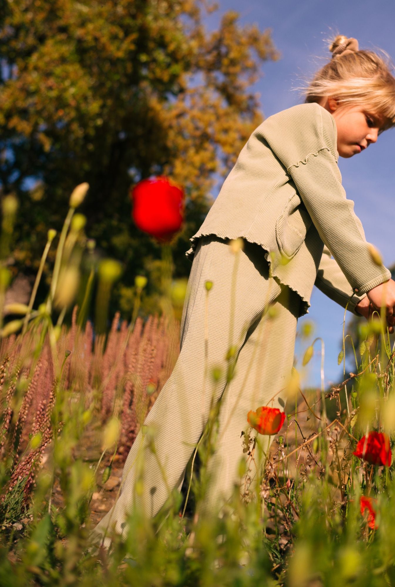 A little girl in the field smelling flowers. She is wearing a comfortable green set (pants and top) in organic cotton waffle.