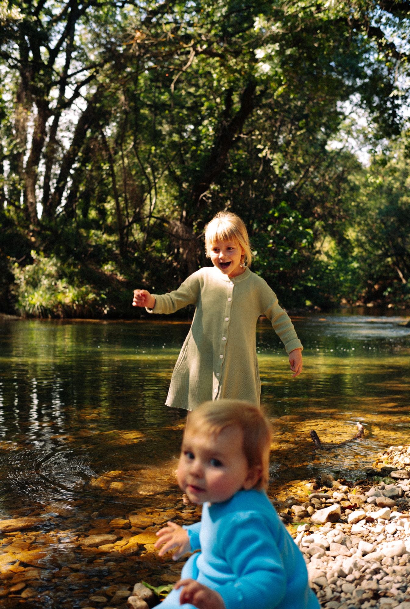 Two sisters playing by the river. Older sister is wearing a long sleeve dress in greenstone. It's made from 100% organic cotton waffle and garment dyed in natural minerals. The younger girl (15 months old) is wearing a sky blue long sleeve dress made from 100% organic cotton.