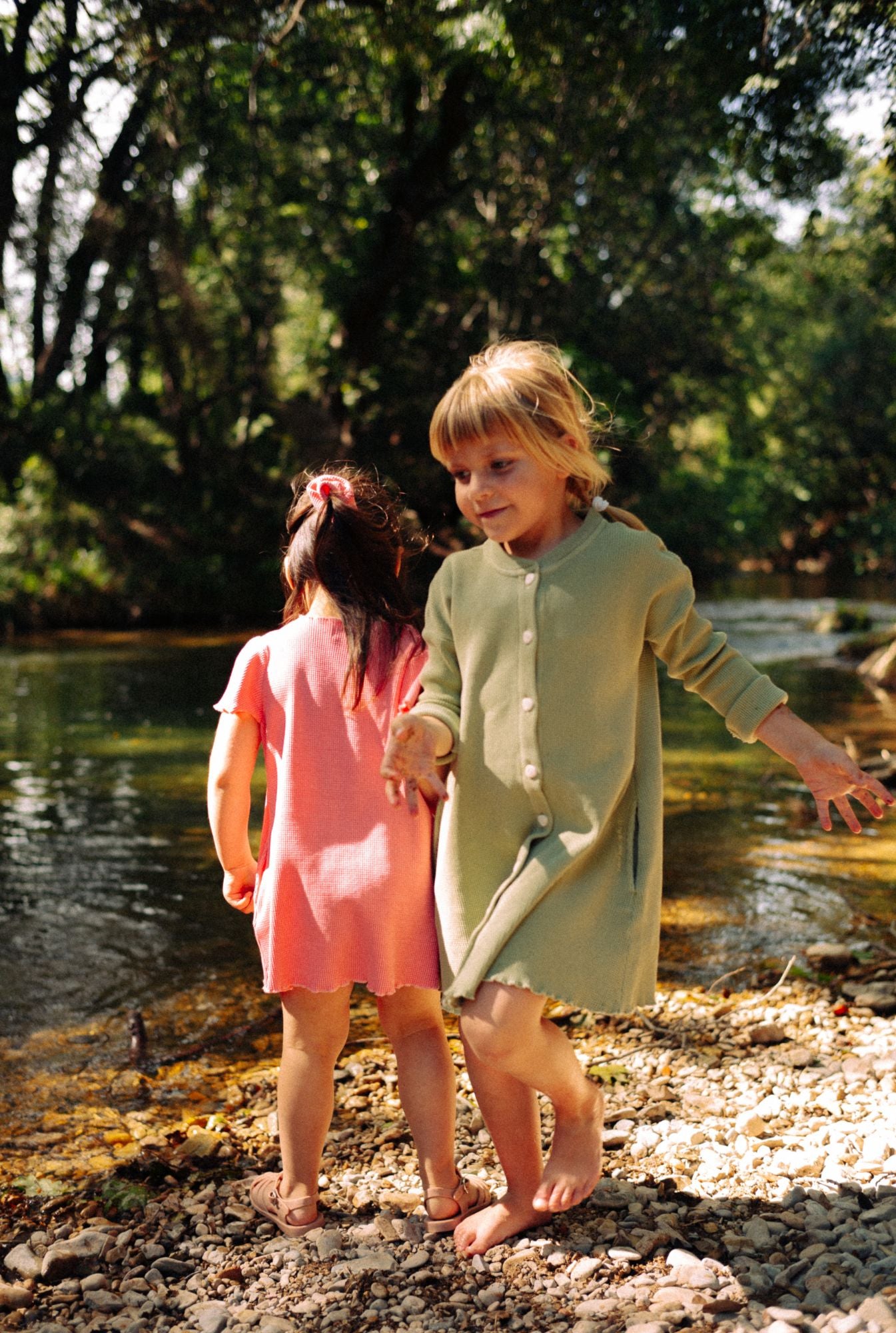 Two little girls are playing by the river. Older is wearing a long sleeve button down dress with pockets and younger is wearing a short sleeve dress in pink. Both are made from 100% organic cotton. They are breathable, comfortable and fun for everyday play.