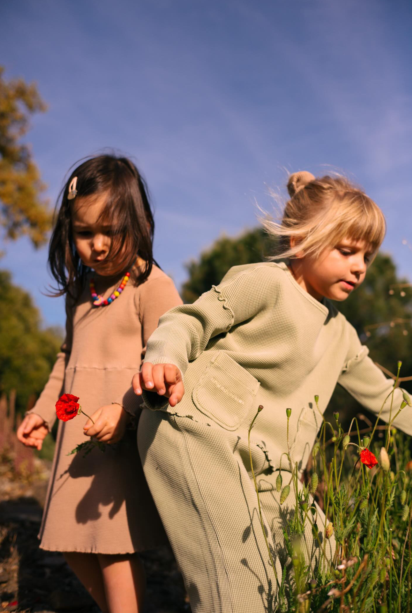 Two girls outside playing. One is wearing long sleeve dress with pockets. The older girl is wearing green top with pants made from organic cotton.