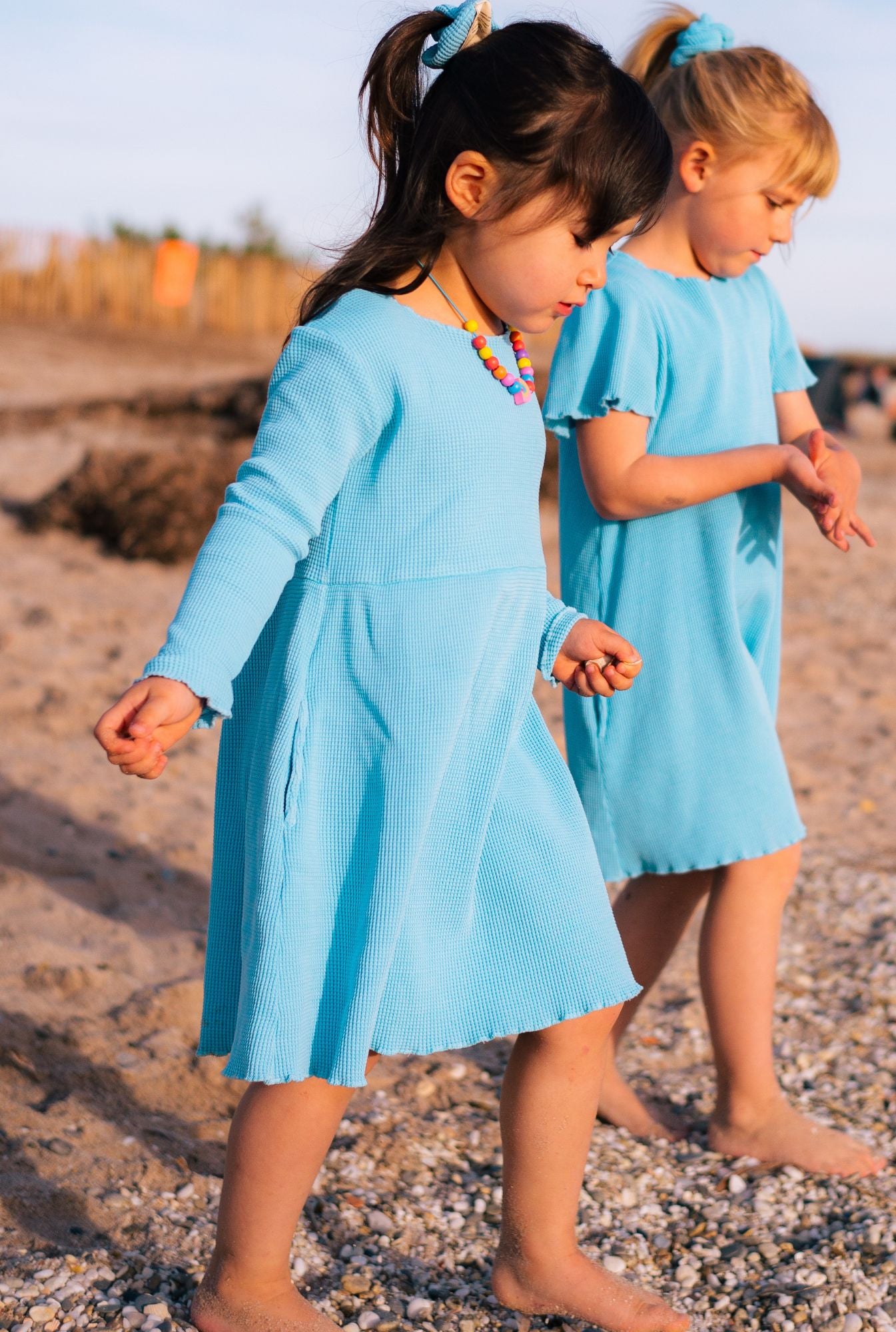 Two girls playing by the beach. One is wearing short sleeve cotton dress in blue. The younger (3 years old) is wearing long sleeves dress in blue. Both dresses are made from organic cotton, garment dyed in natural minerals. They are comfortable, colourful, and breathable.