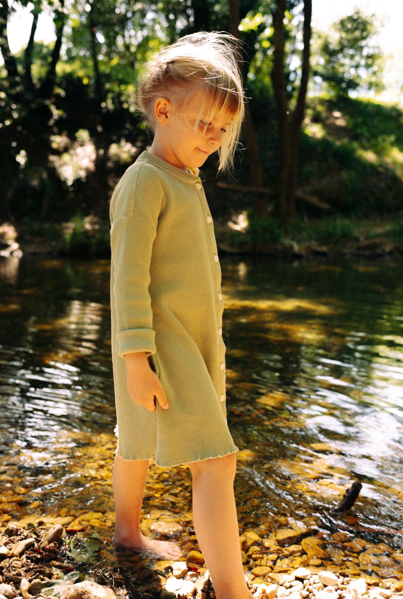 A little girl is playing by the river in a beautiful greenstone dress made from 100% organic cotton waffle and garment dyed in natural minerals.