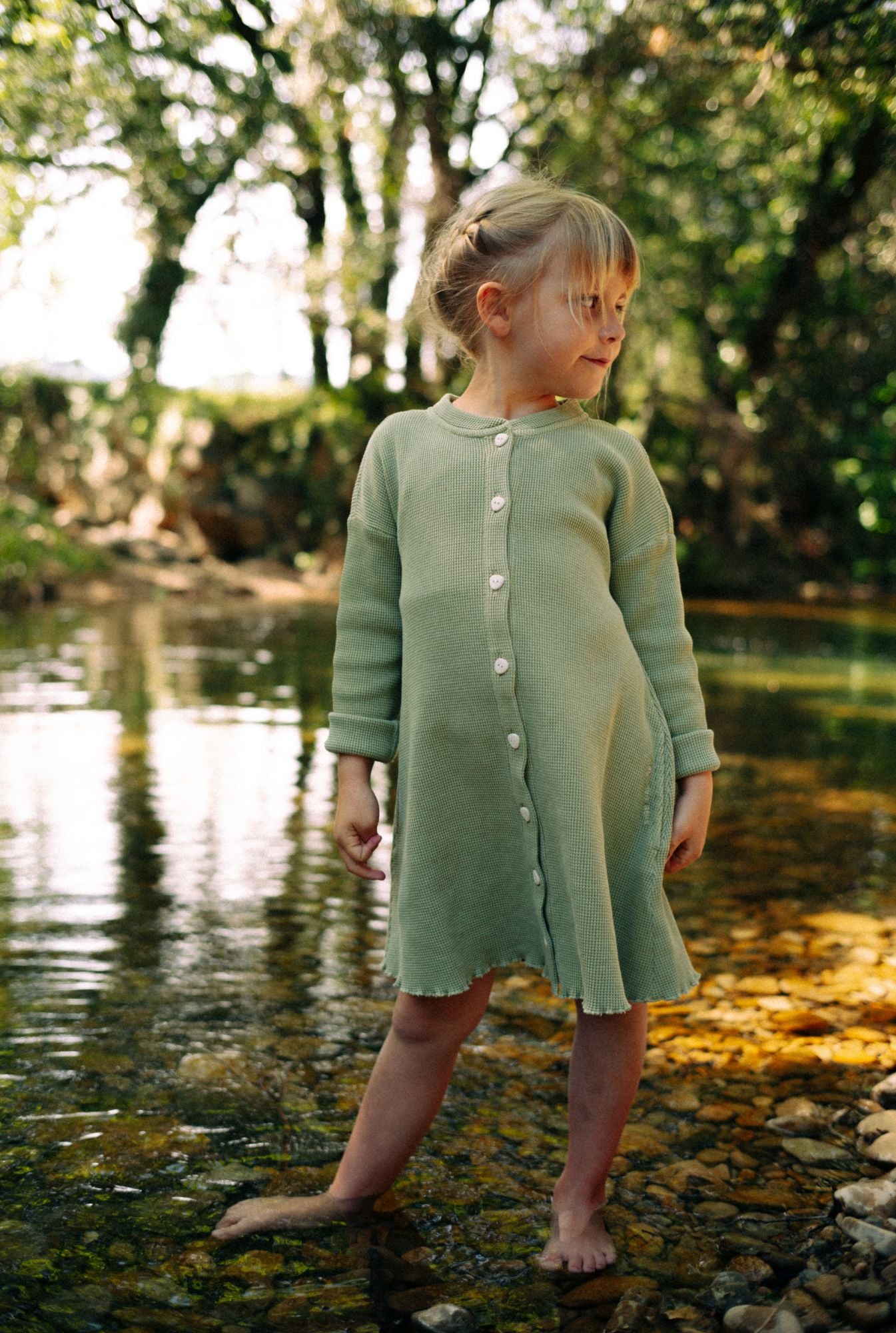 A little girl is playing by the river in a beautiful greenstone dress made from 100% organic cotton waffle and garment dyed in natural minerals.