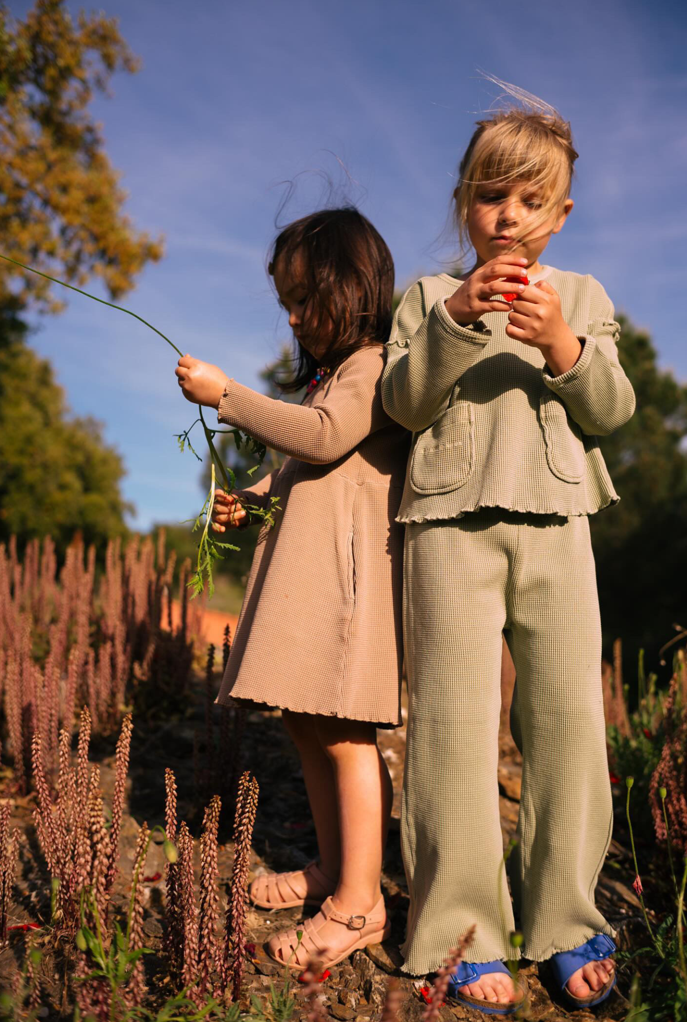 Two little girls playing in nature. Older one is wearing a really comfortable greenstone set and younger one is wearing long sleeve dress with pockets in ochre. The clothes are breathable and made with 100% organic cotton waffle.