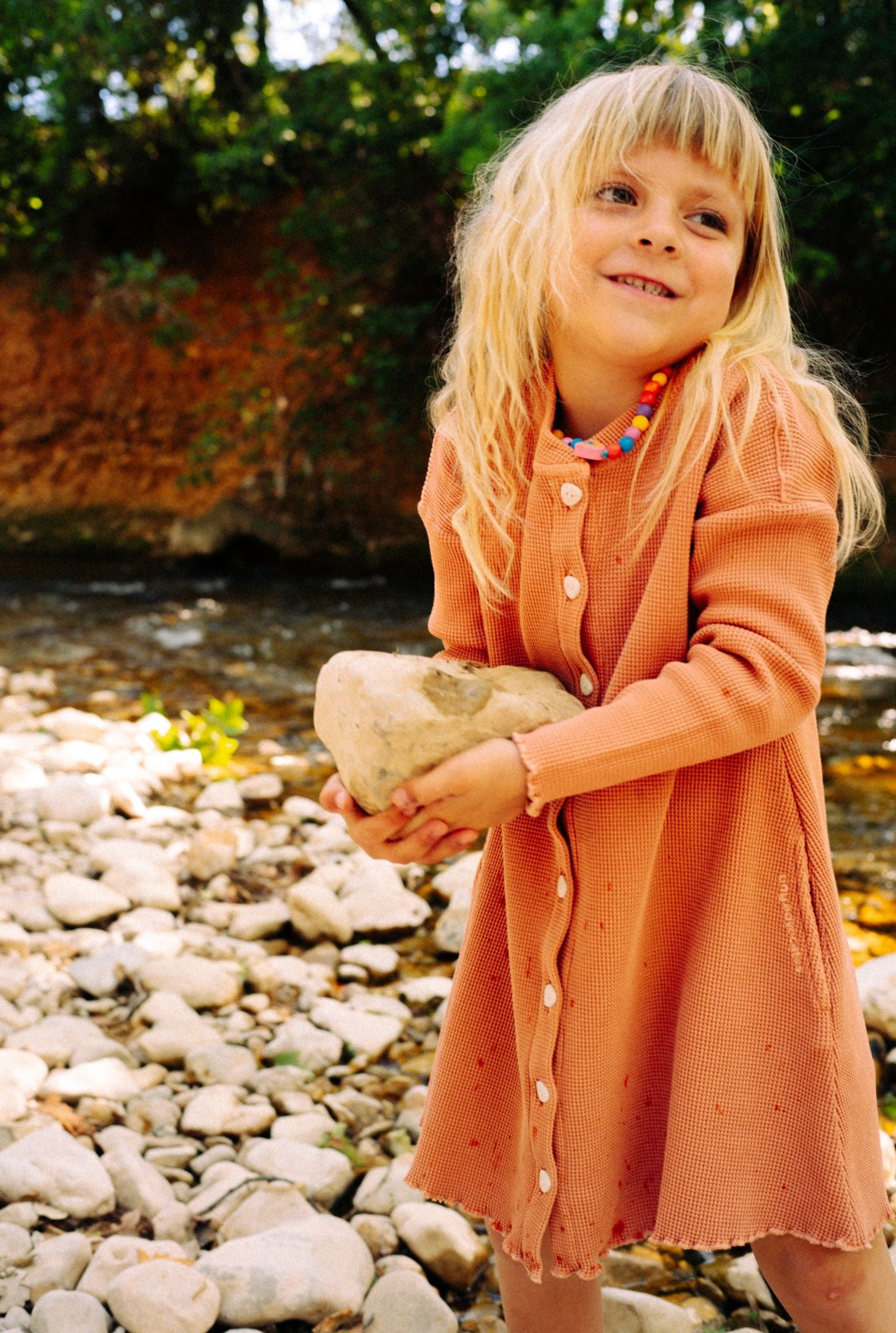 A little girl playing in the river getting splashed by water. She is wearing a comfortable dusty rose dress. The dress is made from 100% organic cotton and garment dyed in natural minerals. It is breathable, comfortable, and fun for everyday play.