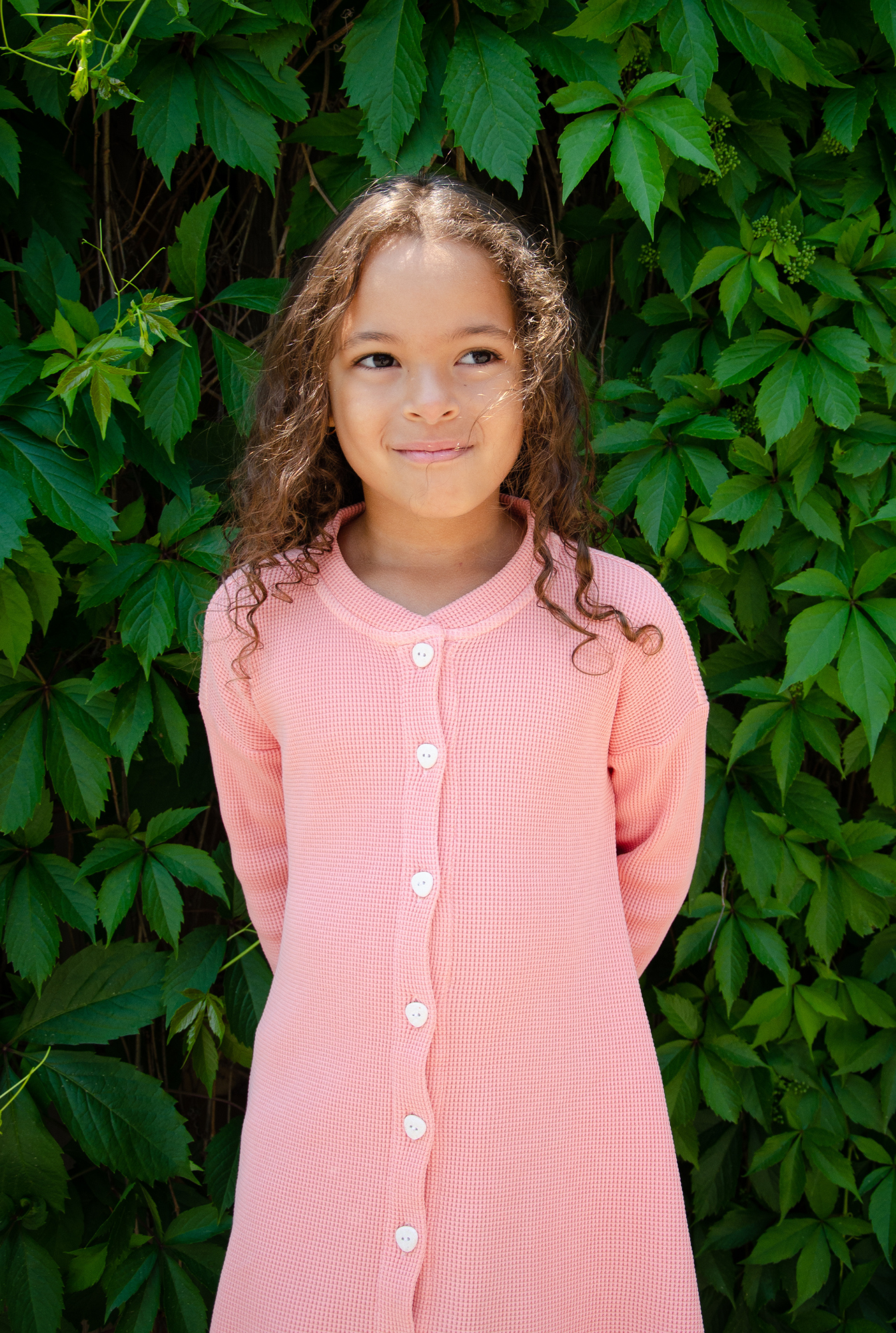 A little girl is wearing a beautiful long sleeve pink dress in the garden. The dress is made from 100% organic cotton and garment d yed in natural minerals. It is comfortable and breathable and perfect for indoor and outdoor play.