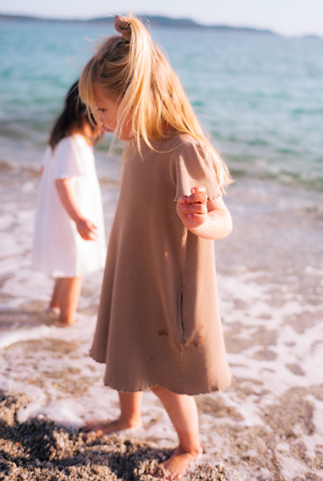 Little girls in summer dresses by the beach. Both wearing short sleeve dresses with pockets. They are made from organic cotton and garment dyed with natural mineral pigments.