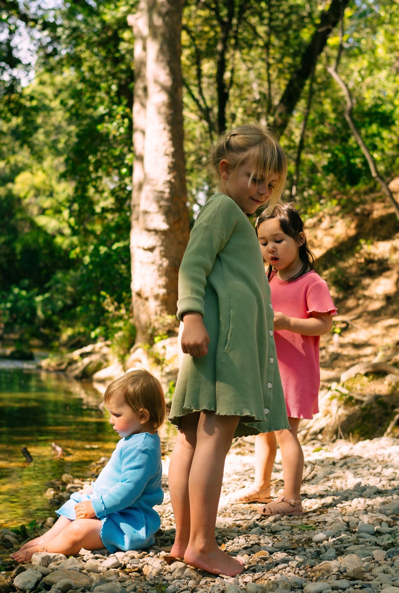 Three little girls are playing by the river They are wearing dresses in green, pink and blue. All made 100% from 100% organic cotton and garment dyed in natural minerals. Comfortable, breathable, and fun for everyday play.