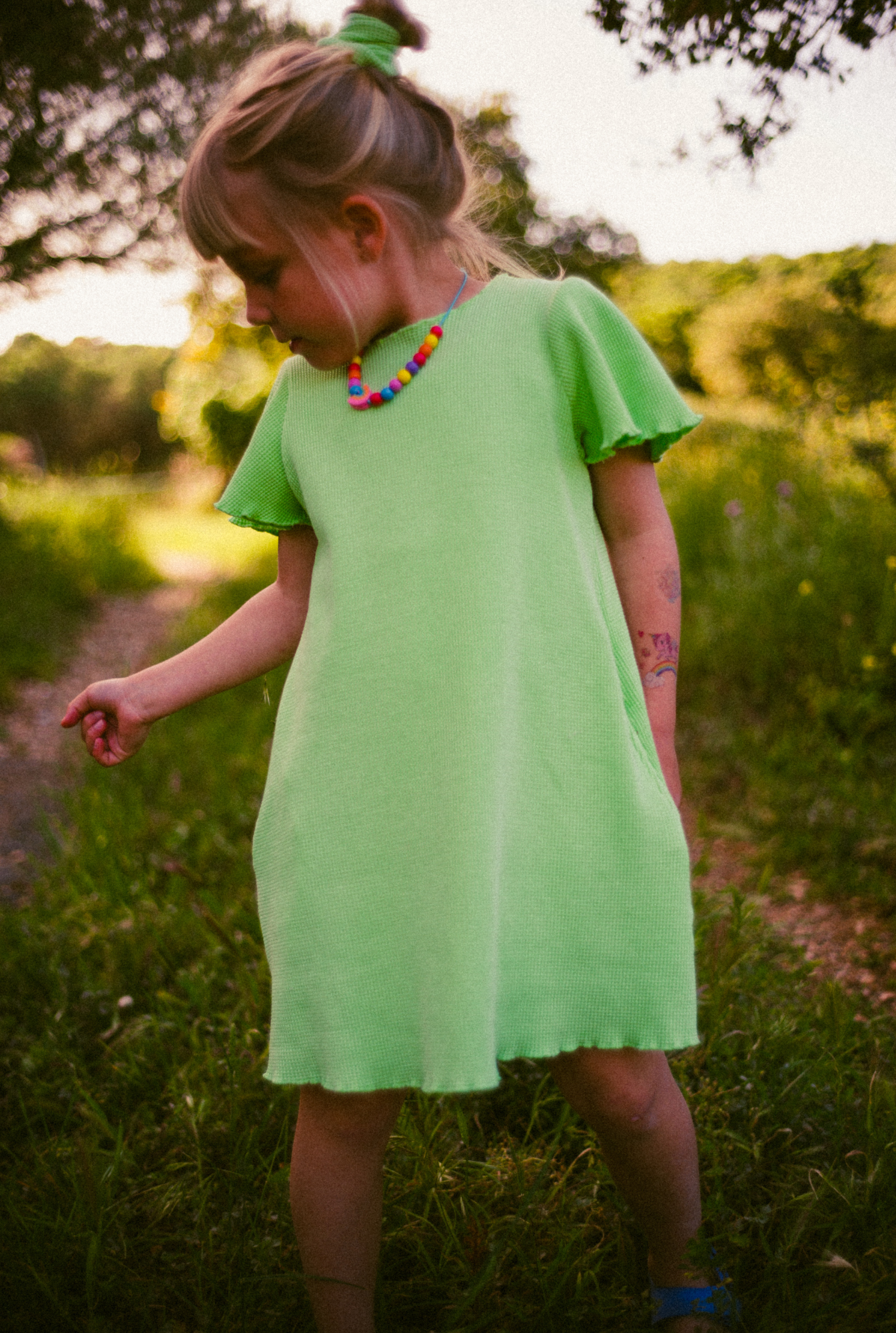 A little girl playing in the field in South of France. She is wearing a beautiful light green dress made from 100% organic cotton. The dress is soft, comfortable and comes with side pockets. matching scrunchie and colourful eco friendly necklace make her outfit even cuter.
