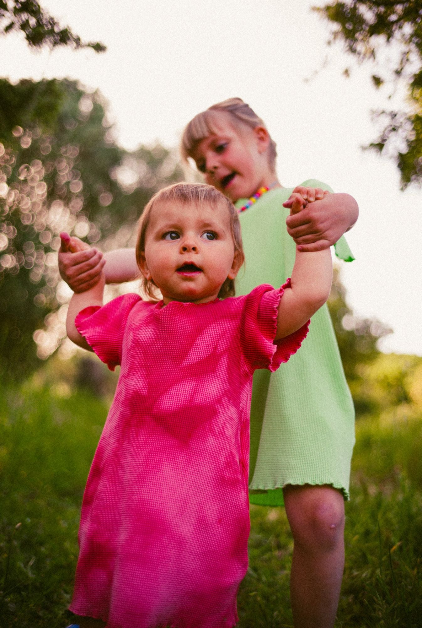 Two little girls in the field wearing short sleeve organic cotton dresses. One is wearing a rose tie dye and the older girl is wearing in light green color.