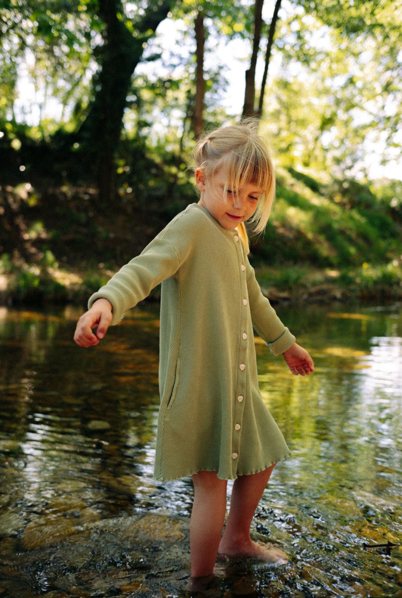 A young girl (6 years old) is playing by the river in South of France. She is wearing a comfortable greenstone dress. It is long sleeves and comes with eco friendly buttons. The dress is breathable and comfortable, made from 100% organic cototn waffle and garment dyed in natural minerals.