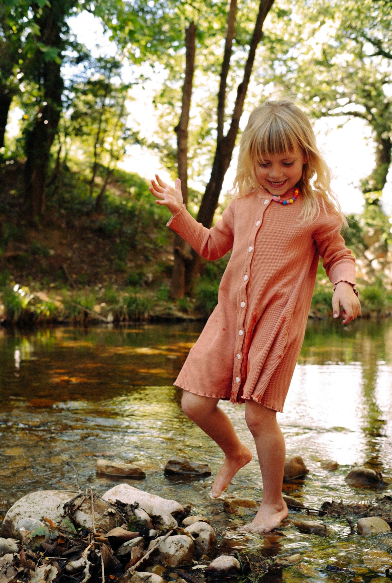 A little girl playing in the river getting splashed by water. She is wearing a comfortable dusty rose dress. The dress is made from 100% organic cotton and garment dyed in natural minerals. It is breathable, comfortable, and fun for everyday play.