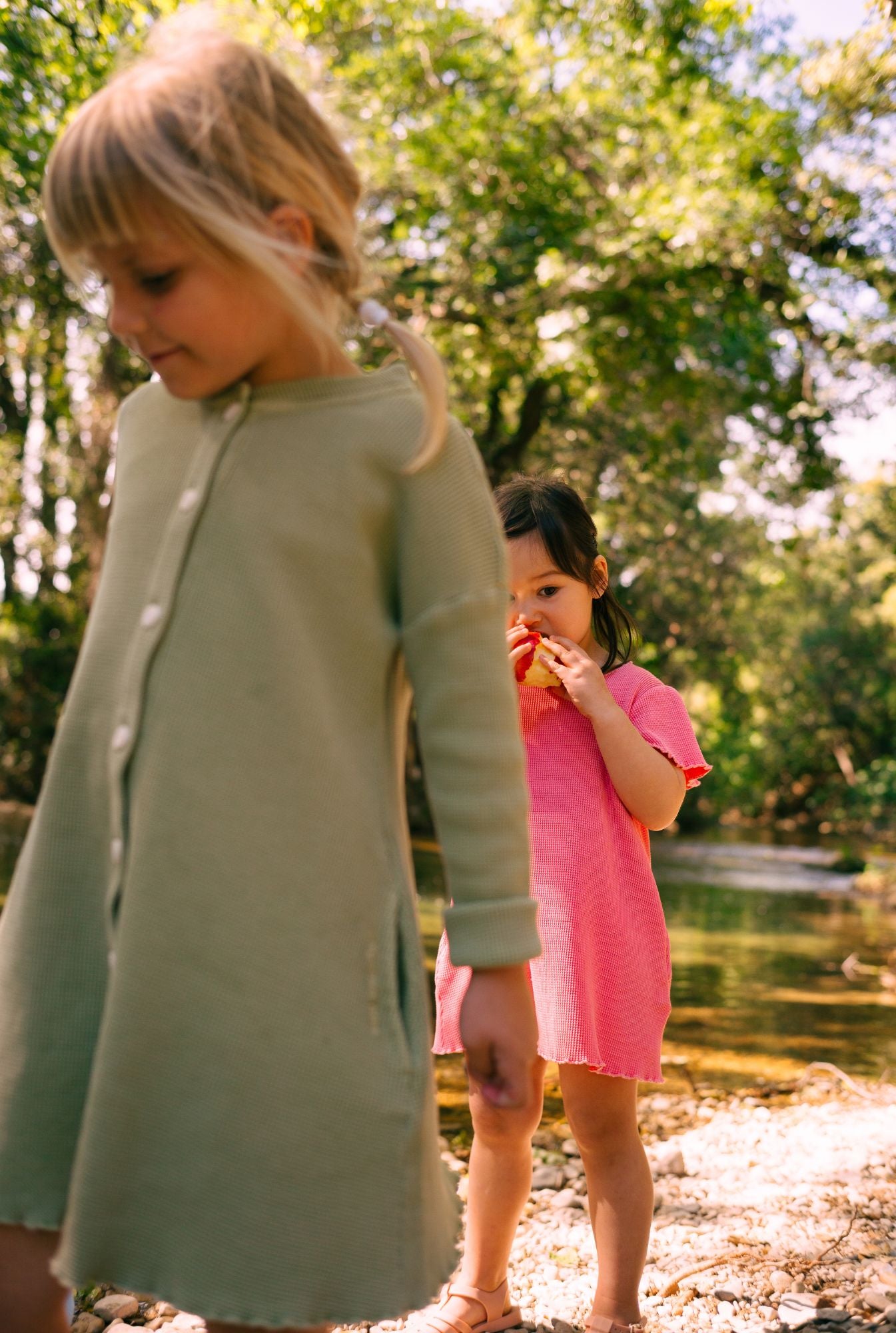 Two little girls are playing by the river. Older is wearing a long sleeve button down dress with pockets and younger is wearing a short sleeve dress in pink. Both are made from 100% organic cotton. They are breathable, comfortable and fun for everyday play.