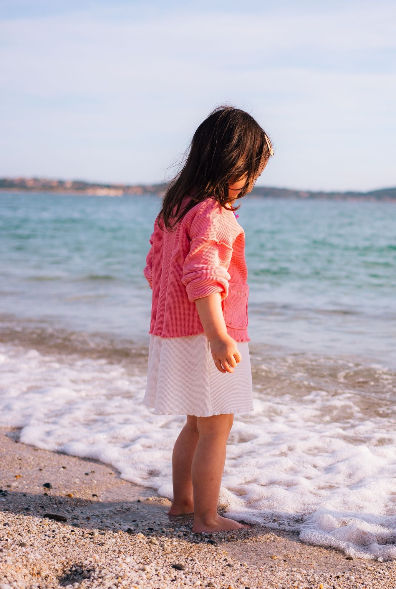 A little girl playing on the beach. She is wearing a beautiful and comfortable long sleeve top in pink and a white dress made from organic cotton waffle.