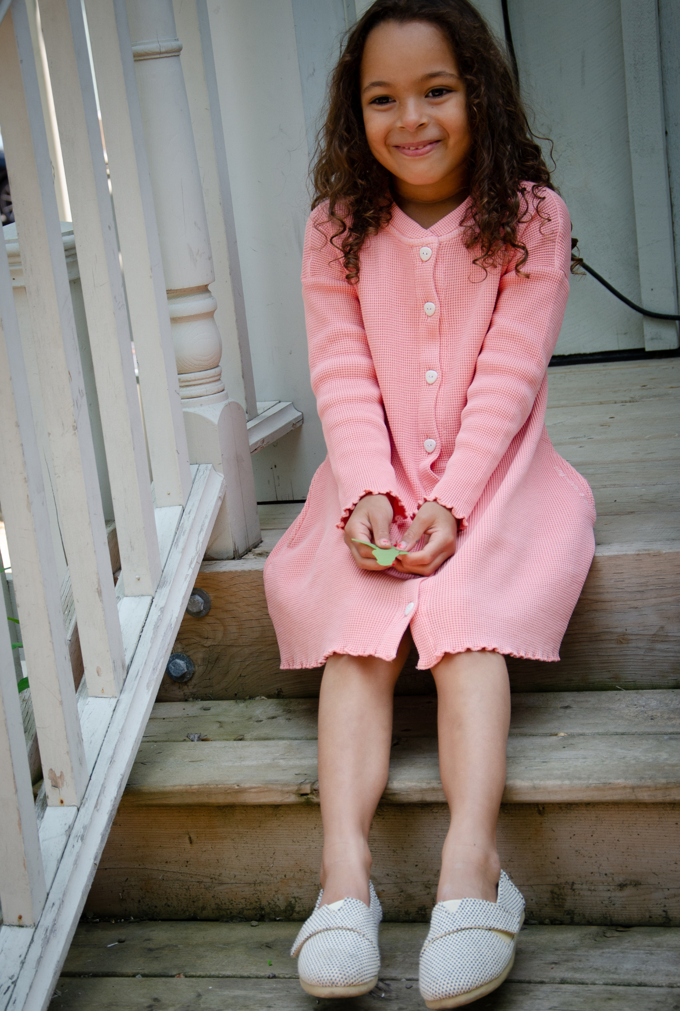 A six years old girl sitting on the staircase outside. She is wearing a beautiful long sleeve pink dress. Its made from 100% organic cotton and garment dyed in natural minerals. It's breathable and can be worn all year round.