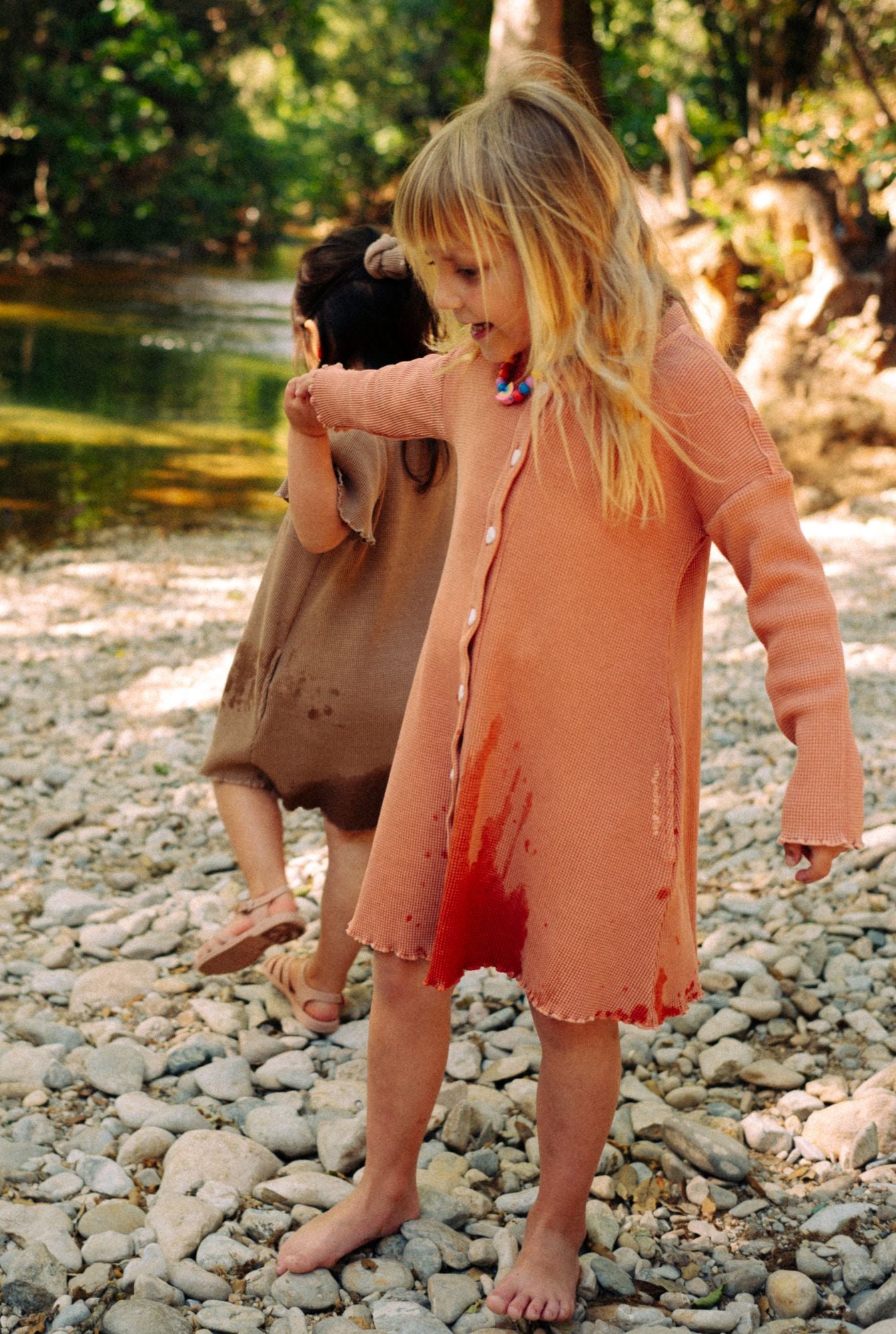 Two little girls are playing by the river in their comfy 100% organic cotton waffle dresses. One is wearing a long sleeve dress in dusty rose with buttons. The other girl is wearing short sleeve ochre dress with pockets. These dresses are breathable and fun for everyday play.