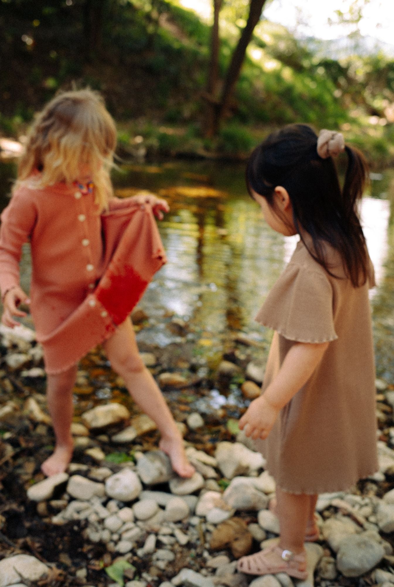 Two little girls are playing by the river in their comfy 100% organic cotton waffle dresses. One is wearing a long sleeve dress in dusty rose with buttons. The other girl is wearing short sleeve ochre dress with pockets. These dresses are breathable and fun for everyday play.