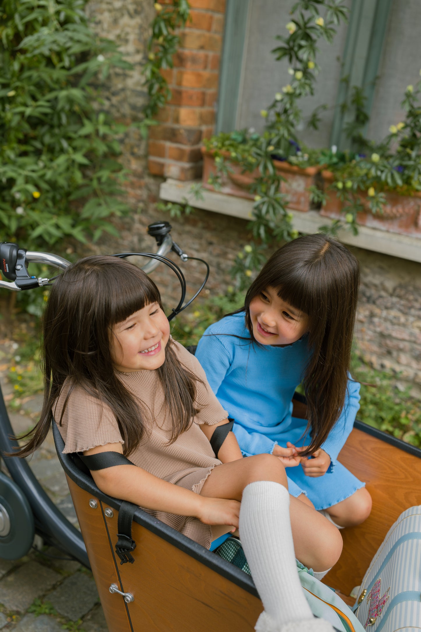 Two little girls are wearing colourful organic cotton dresses on their way to school. One is wearing long sleeve dress in blue and the other is wearing short sleeve dress in ochre.