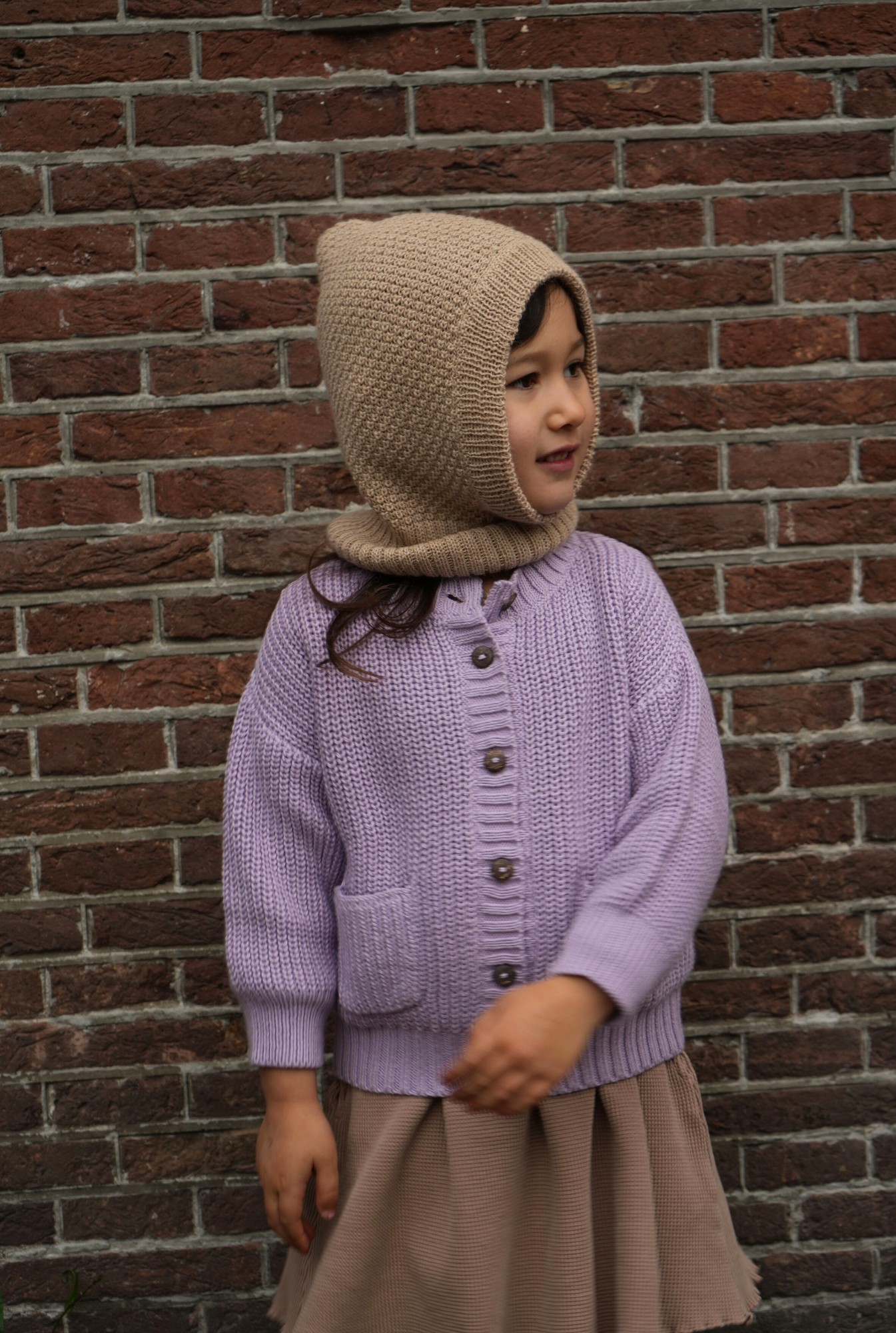 Child wearing a purple knitted cardigan and beige baby alpaca hat against a brick wall