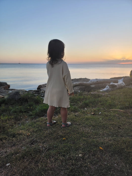 A toddle standing by the ocean and looking at sunset. She is wearing a beautiful organic cotton yellow dress with long sleeves and pockets. 