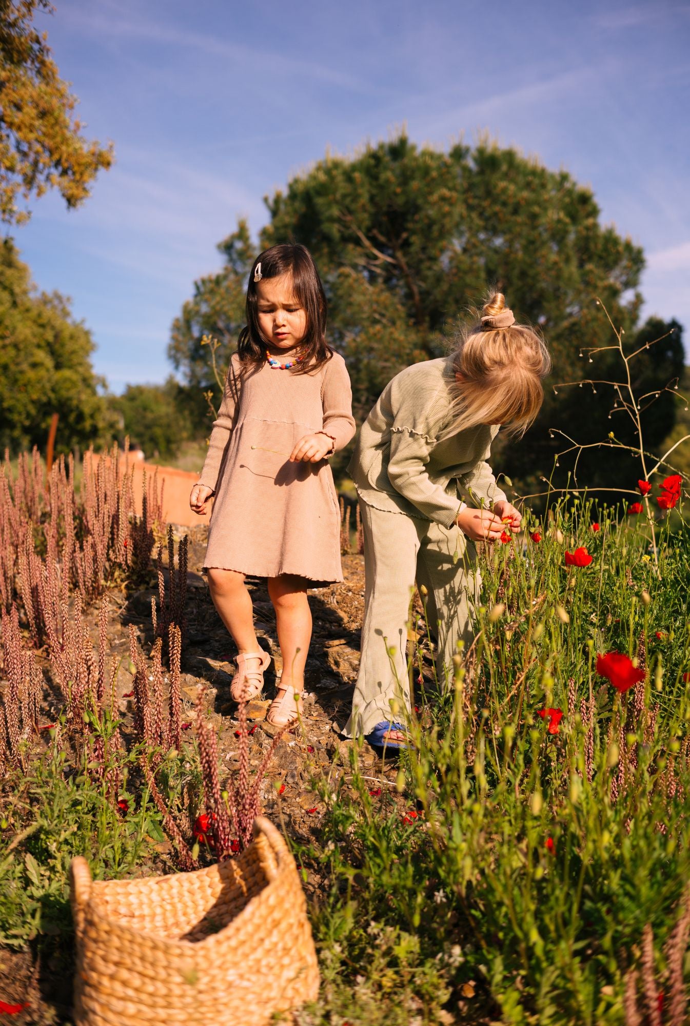 Two little girls playing in nature. Older one is wearing a really comfortable greenstone set and younger one is wearing long sleeve dress with pockets in ochre. The clothes are breathable and made with 100% organic cotton waffle.