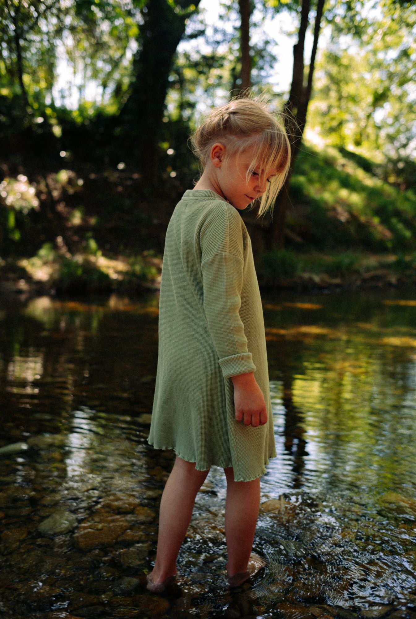 A little girl is playing by the river in a beautiful greenstone dress made from 100% organic cotton waffle and garment dyed in natural minerals.