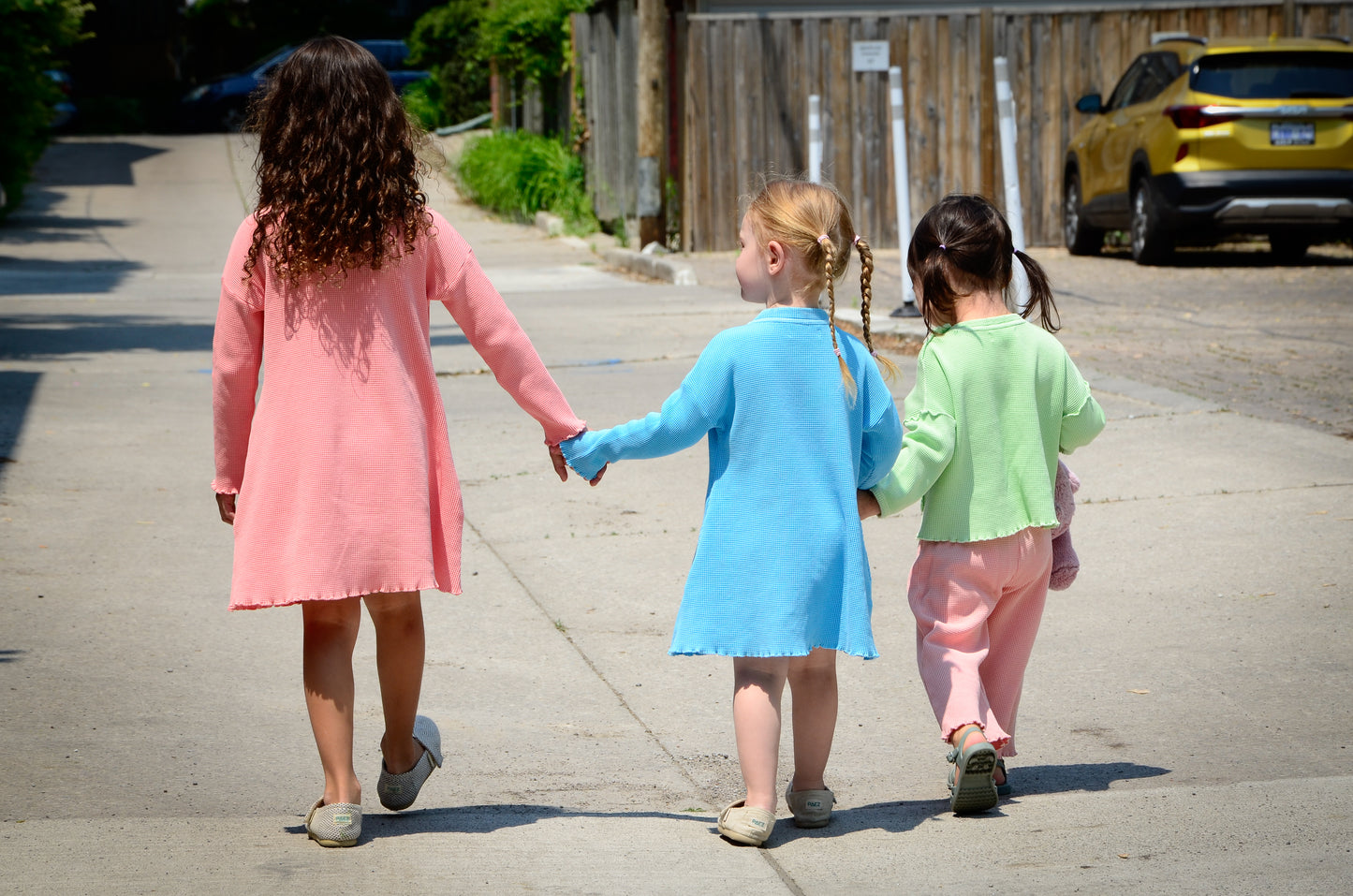 3 kids walking down holding hands. Older girl is wearing a long sleeve dress with pockets. Another girl is wearing a beautiful sky blue long sleeve dress with pockets. The youngest girl is wearing a light green long sleeve top and pink wide leg pants. All pieces are made from 100% organic cotton waffle fabric that is breathable for all year round wear.