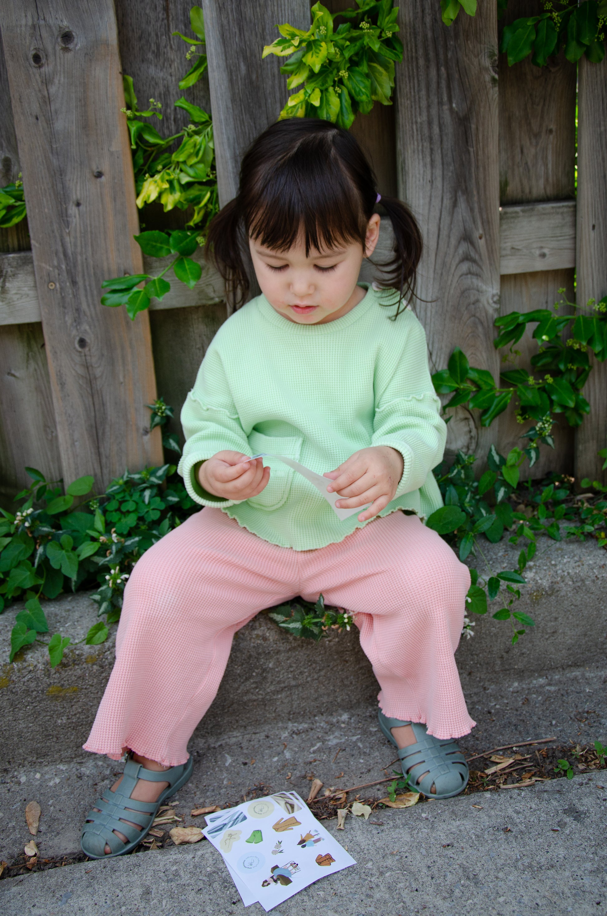 A little girl is playing outside. She is wearing a mix and match set with light green long sleeve top and light pink wide leg pants. Both made from organic cotton.