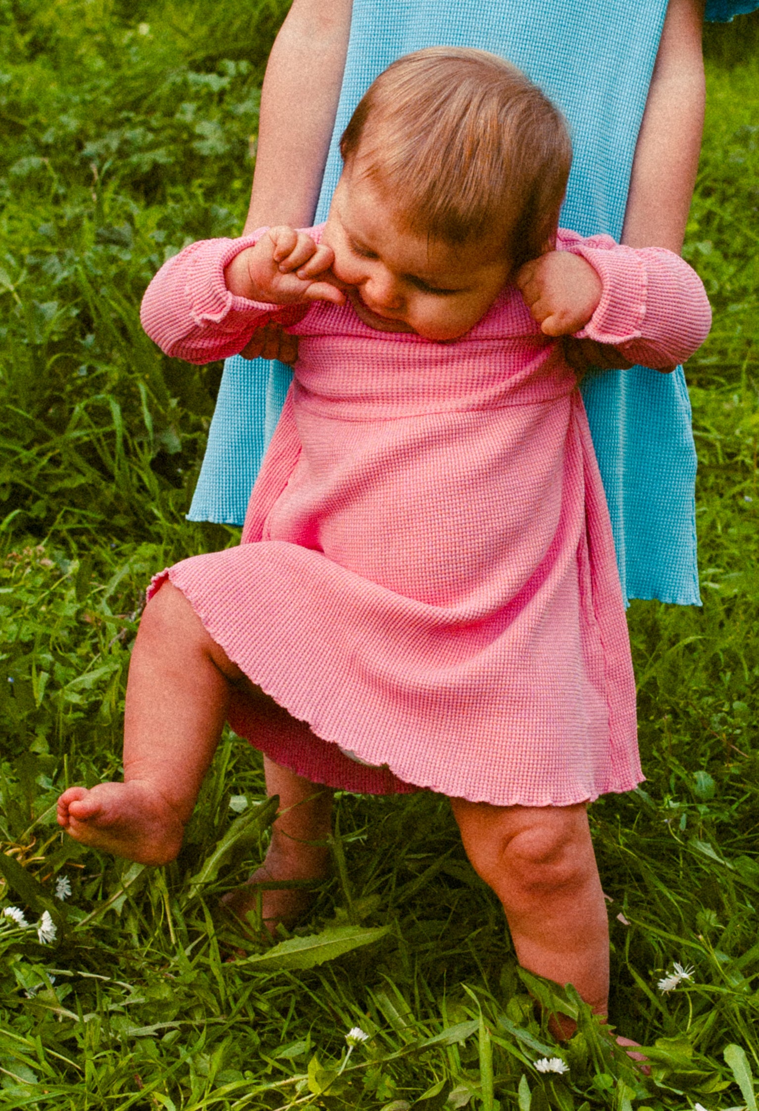 A little girl and her sister in the field. They are both wearing cotton dresses. Young girl is wearing long sleeves with pockets pink cotton dress. Her older sister is wearing short sleeves blue dress with pockets.