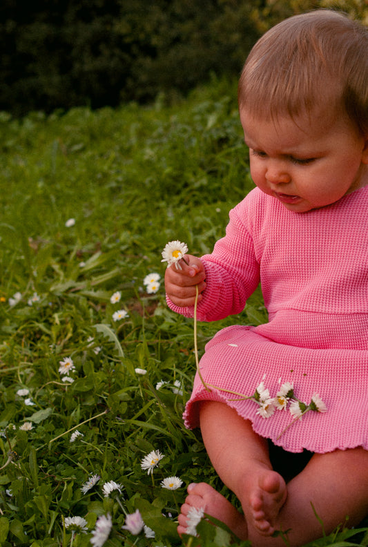 A little girl sitting on the grass playing with flowers. She is wearing a long sleeve pink dress made from 100% organic cotton waffle dress. This beautiful, comfortable, breathable, and affordable dress is crafted for everyday indoor and outdoor play.