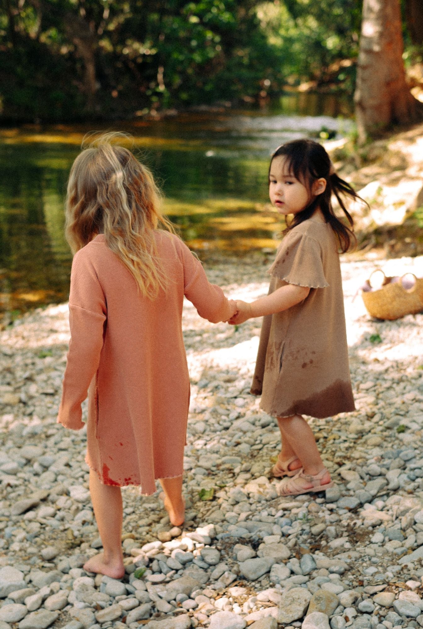 Two little girls are playing by the river in their comfy 100% organic cotton waffle dresses. One is wearing a long sleeve dress in dusty rose with buttons. The other girl is wearing short sleeve ochre dress with pockets. These dresses are breathable and  fun for everyday play.