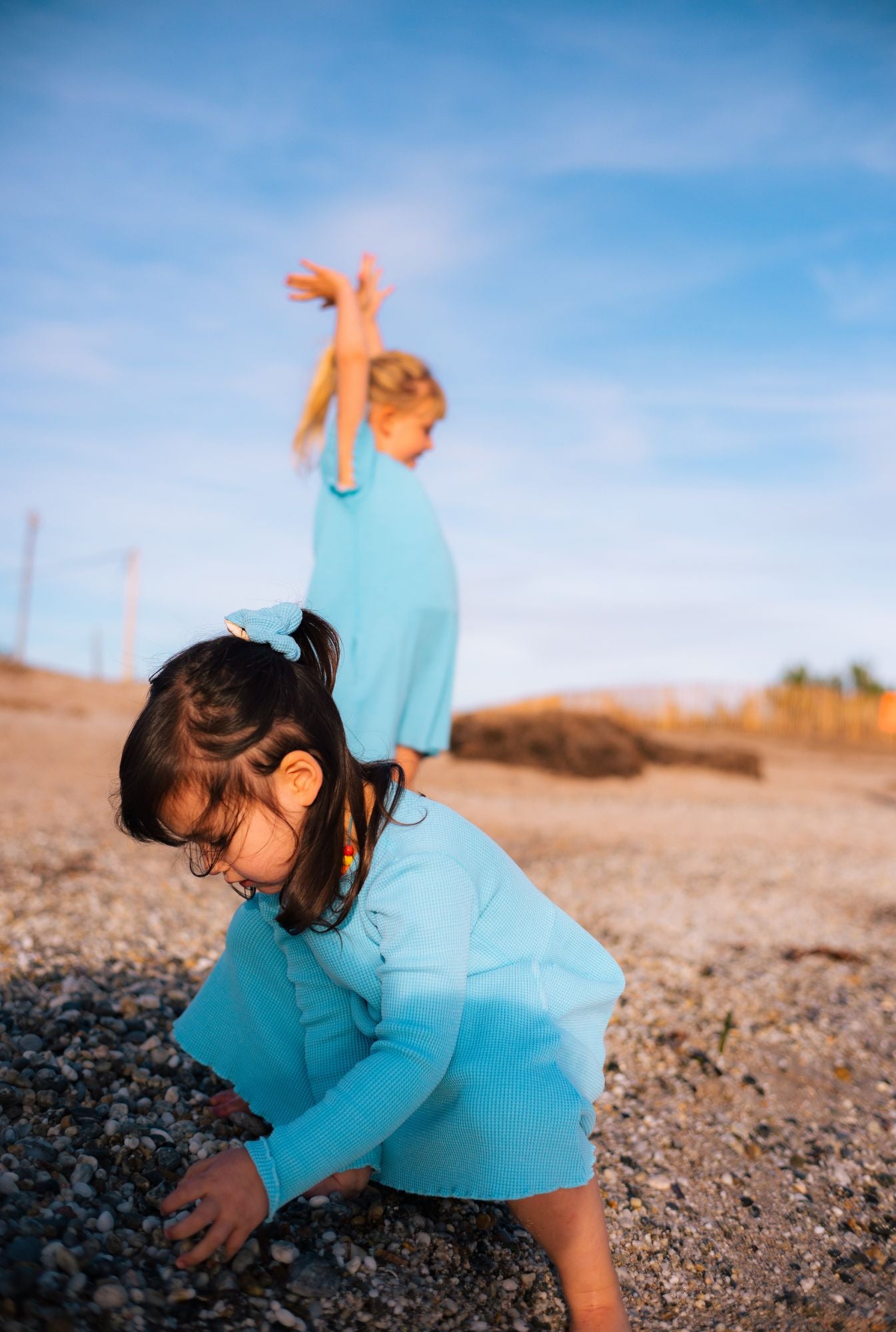 Two girls playing by the beach. Older girl is wearing a short sleeve dress with pockets. The younger girl is wearing a long sleeve dress with pockets in blue. Both dresses are made with 100% organic cotton.