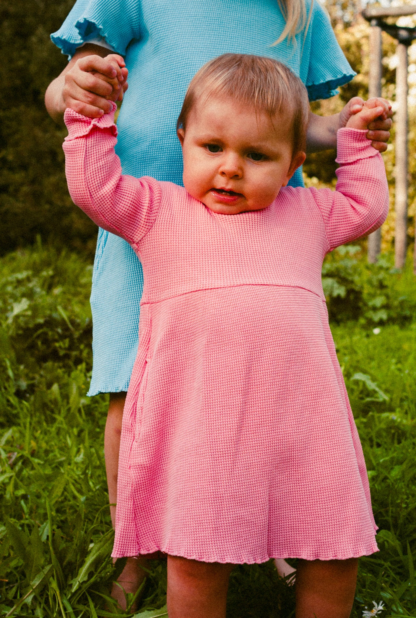 A little girl and her sister in the field. They are both wearing cotton dresses. Young girl is wearing long sleeves with pockets pink cotton dress. Her older sister is wearing short sleeves blue dress with pockets.