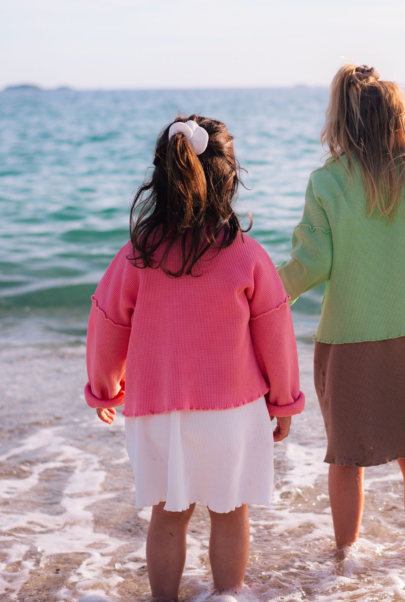 Two kids by the beach playing. They are wearing colourful tops and dresses made from organic cotton by Tiny Advocates. 