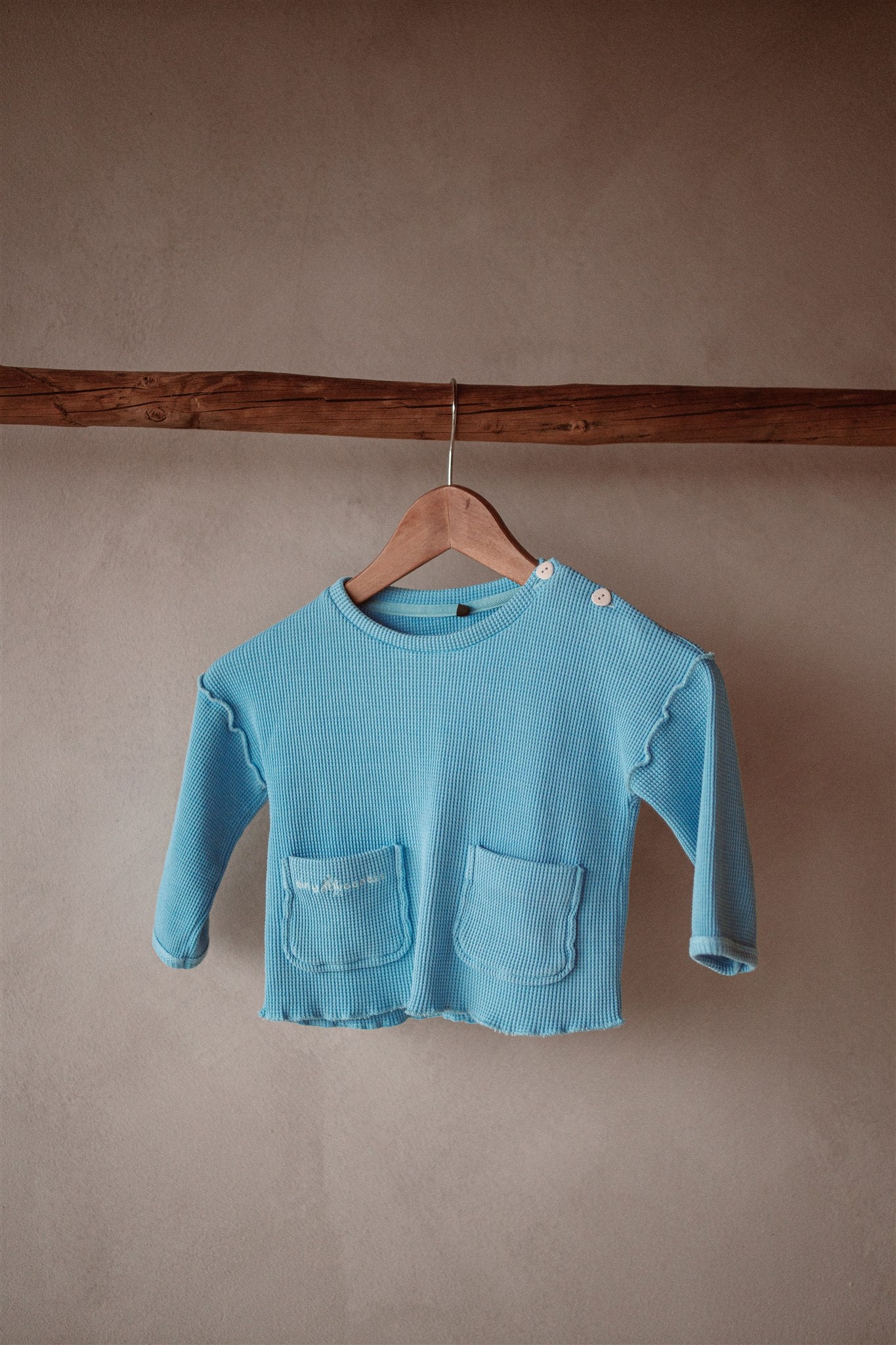 An image of organic cotton kids long sleeve top. It features long sleeves, front pockets, and lettuce hem finish. 