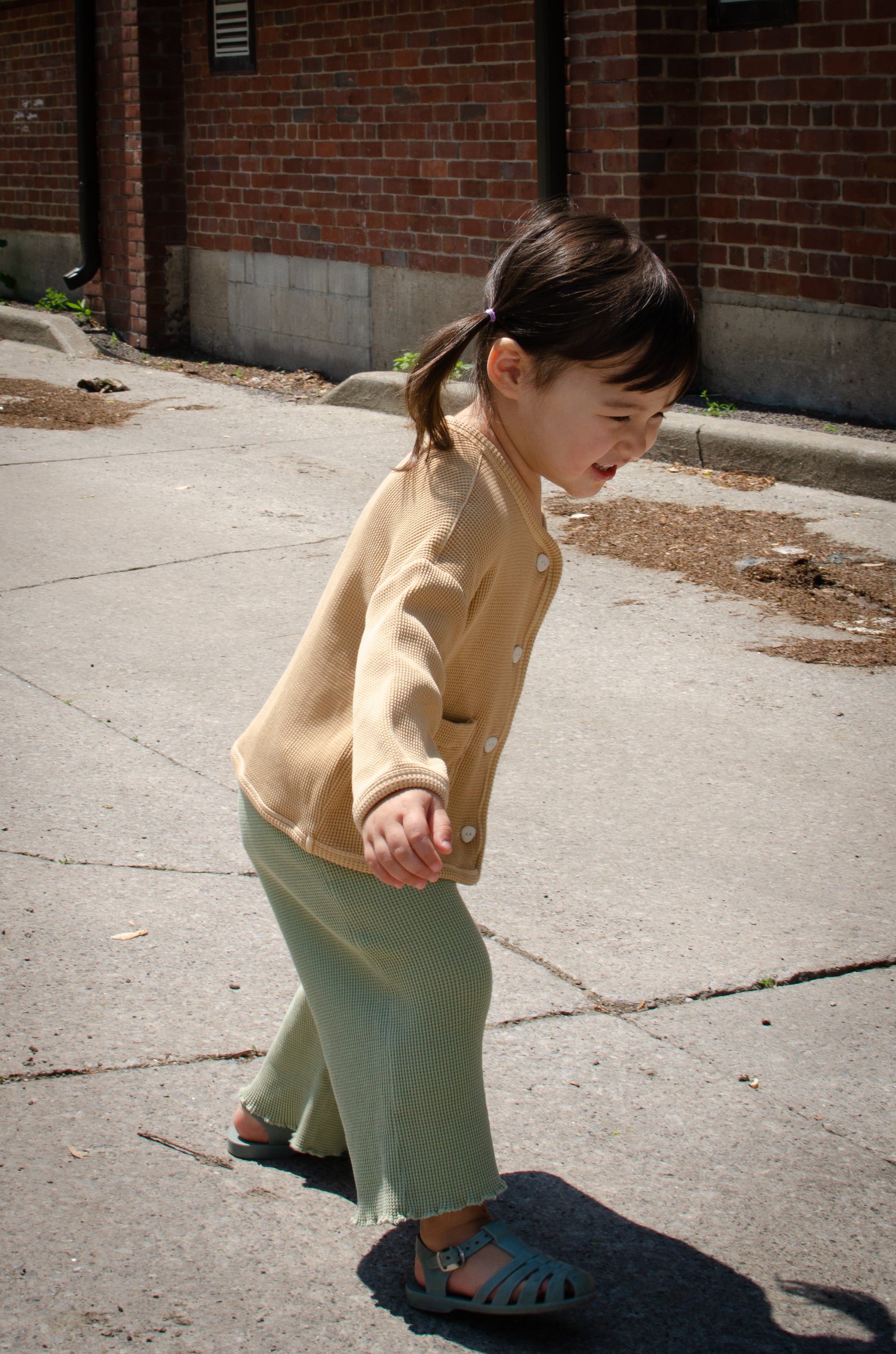 A little girl in the backyard. She is wearing a stylish and comfortable outfit - a sandstone cardigan with eco-friendly buttons and wide leg pants in green made from organic cotton.