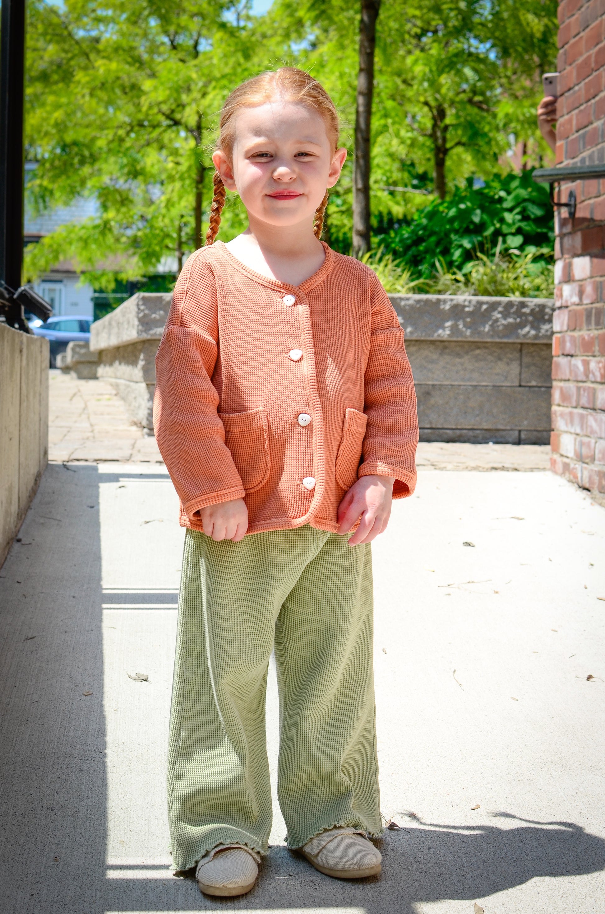 A little girl standing in her stylish and comfortable mix and match set by Tiny Advocates. Wearing a sienna cardigan in size 3T and wide leg greenstone pants with elastic waistband. 