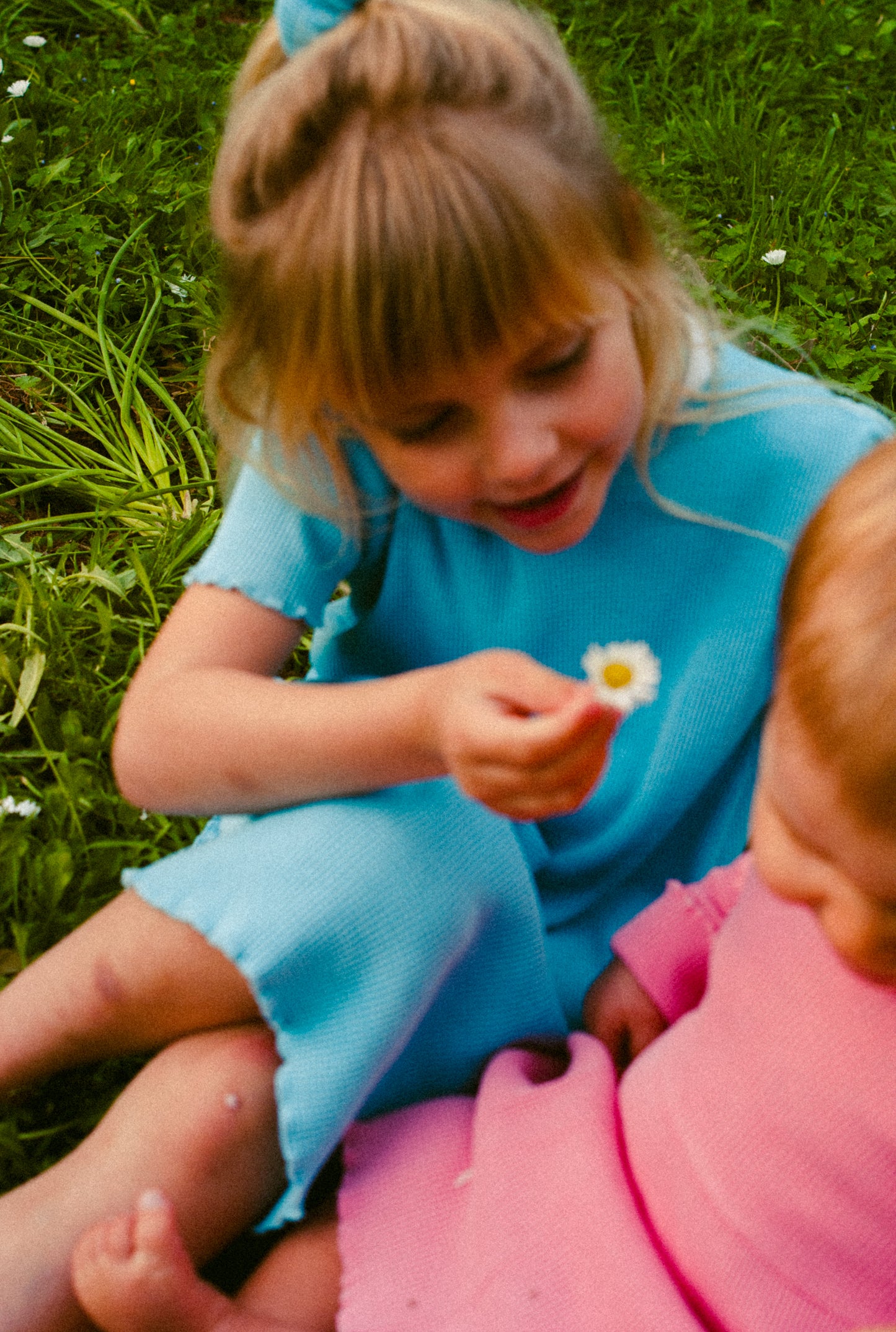 Two little girls in the field smelling flowers. Older one is wearing a short sleeve blue dress and young one is wearing a long sleeve pink dress made from organic cotton waffle. Both dresses have pockets.