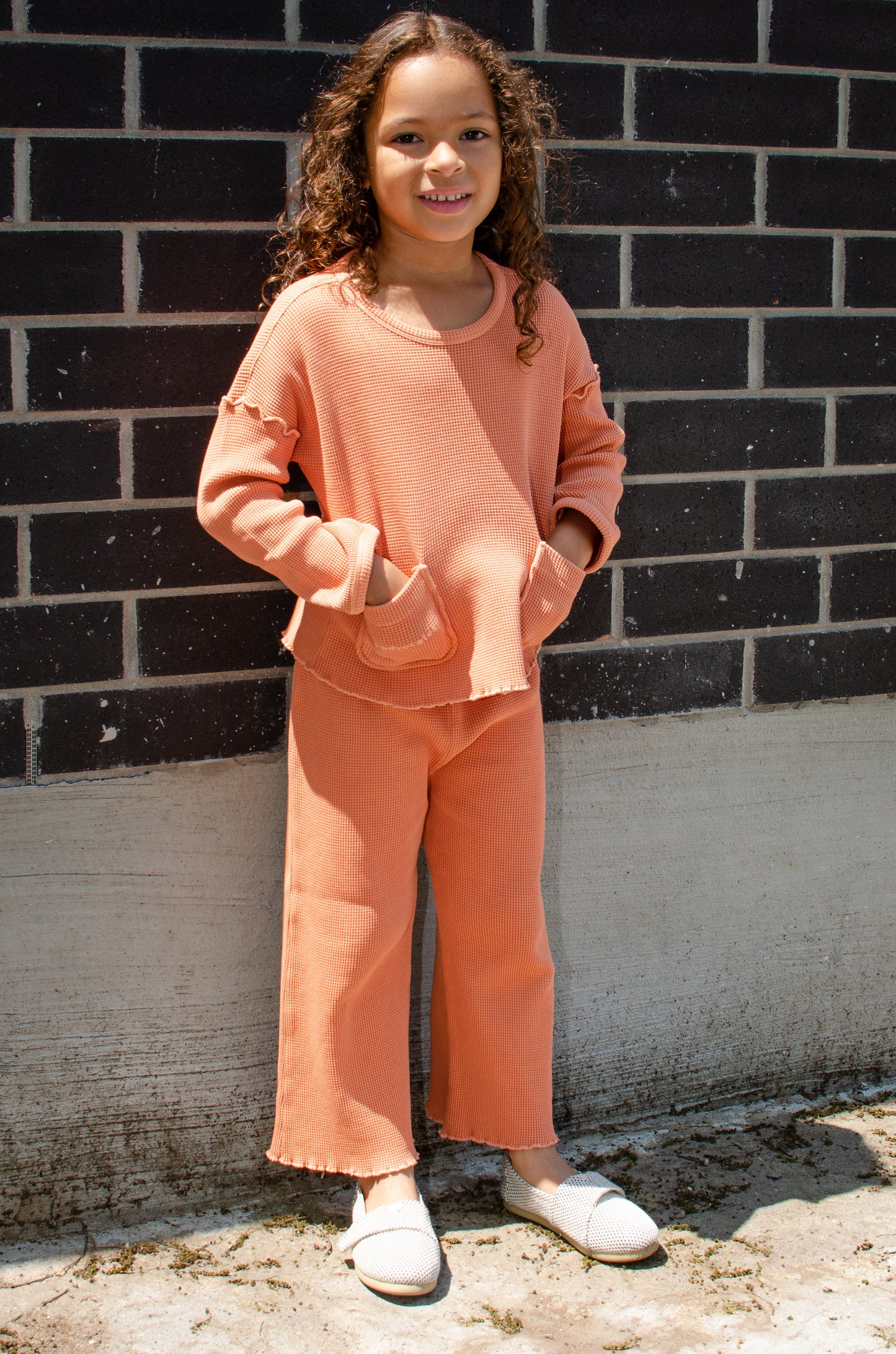 A little girl standing in a beautiful Sienna set (pants and top) made from organic cotton and garment dyed in natural mineral pigments. It looks soft and stylish. The photo taken in Toronto.