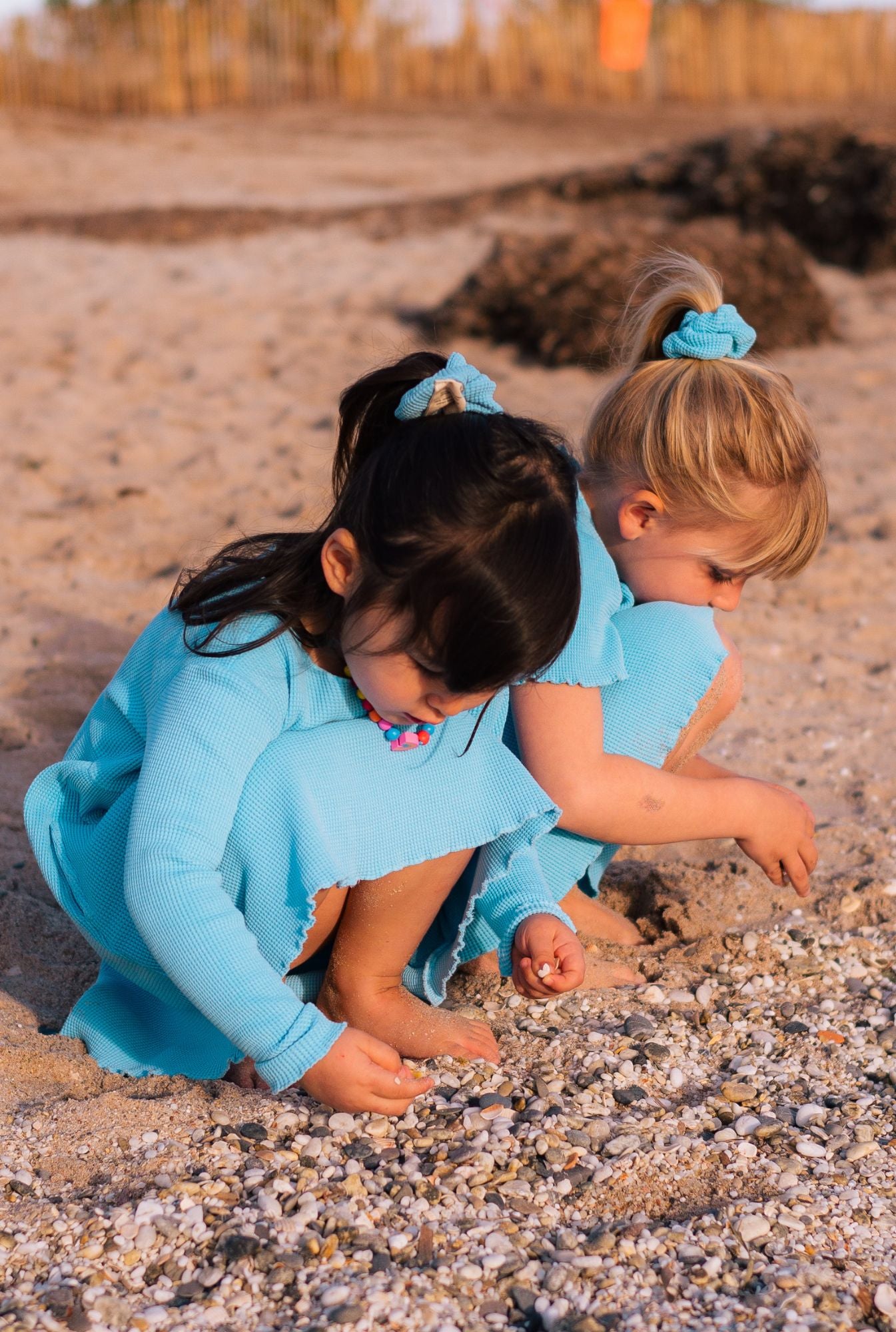 Two girls playing by the beach. One is wearing short sleeve cotton dress in blue. The younger (3 years old)  is wearing long sleeves dress in blue. Both dresses are made from organic cotton, garment dyed in natural minerals. They are comfortable, colourful, and breathable.